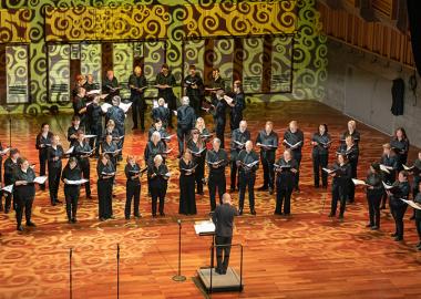 singers on stage standing in a semi-circle, being led by the conductor